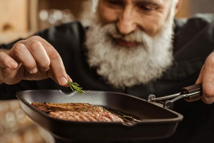 aged man cooking a steak in an iron cast
