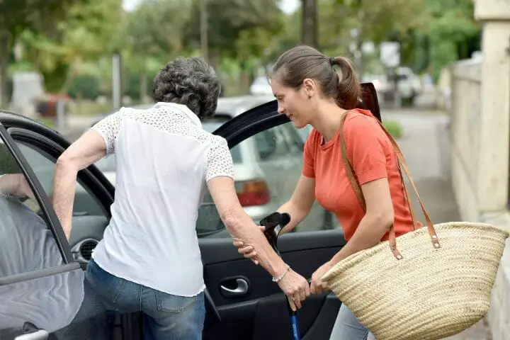 Caregiver helping a senior woman with crane to get in the passenger seat of a car